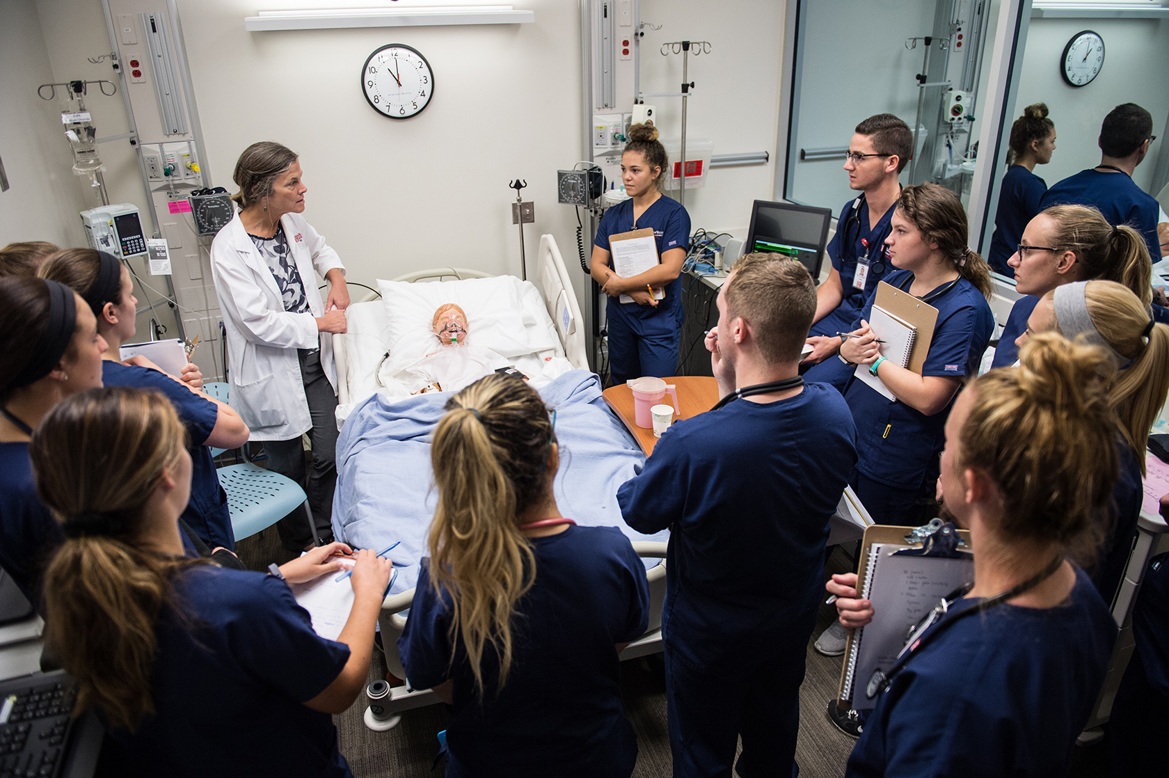 Female in white coat talking with students in blue scrubs at hospital bedside